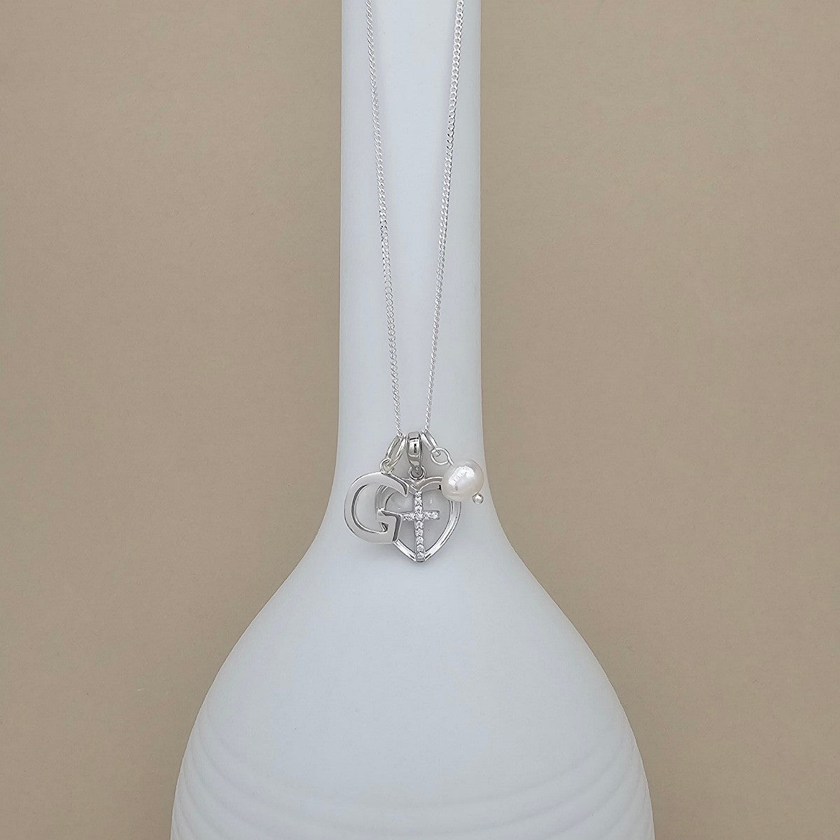 A silver necklace with a heart-shaped pendant and a small pearl accent, displayed on a white ceramic vase against a beige background.