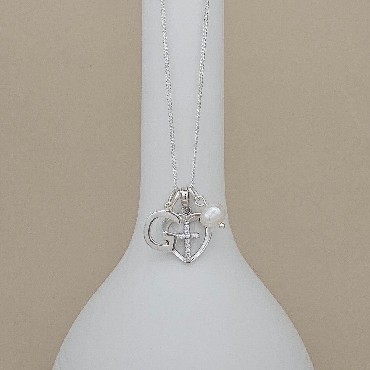 A silver necklace with a heart-shaped pendant featuring a cross and the letter 'G', displayed on a white ceramic vase against a beige background.