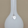 A silver necklace with a heart-shaped pendant engraved with 'Poppy' and a small cross charm, displayed on a white ribbed vase against a beige background.