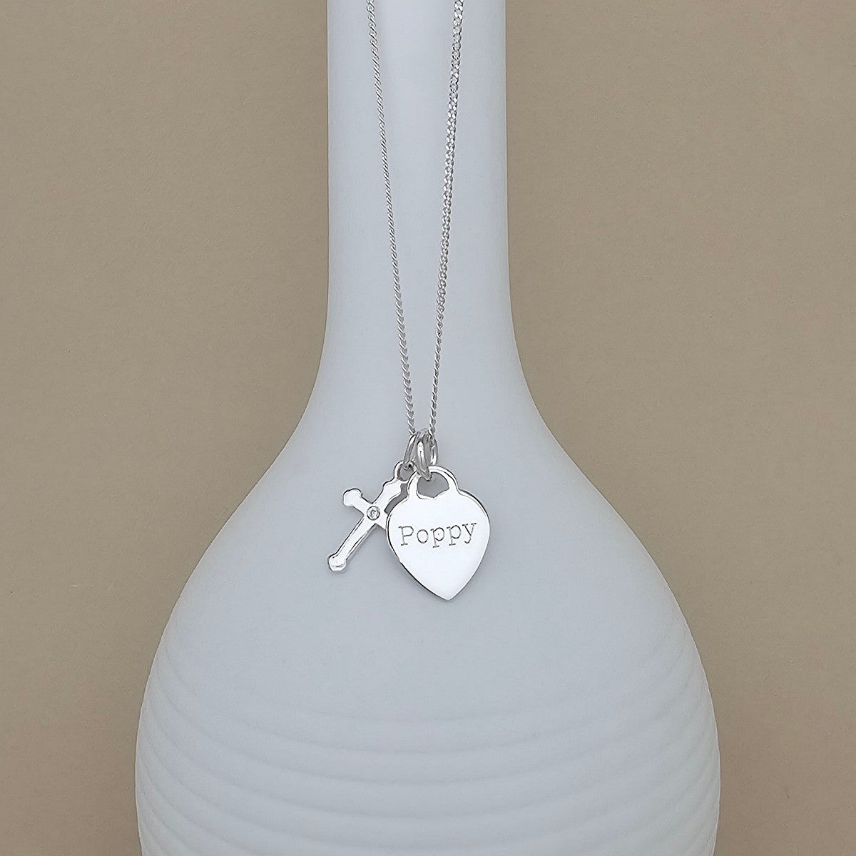 A silver necklace with a heart-shaped pendant engraved with 'Poppy' and a small cross charm, displayed on a white ribbed vase against a beige background.