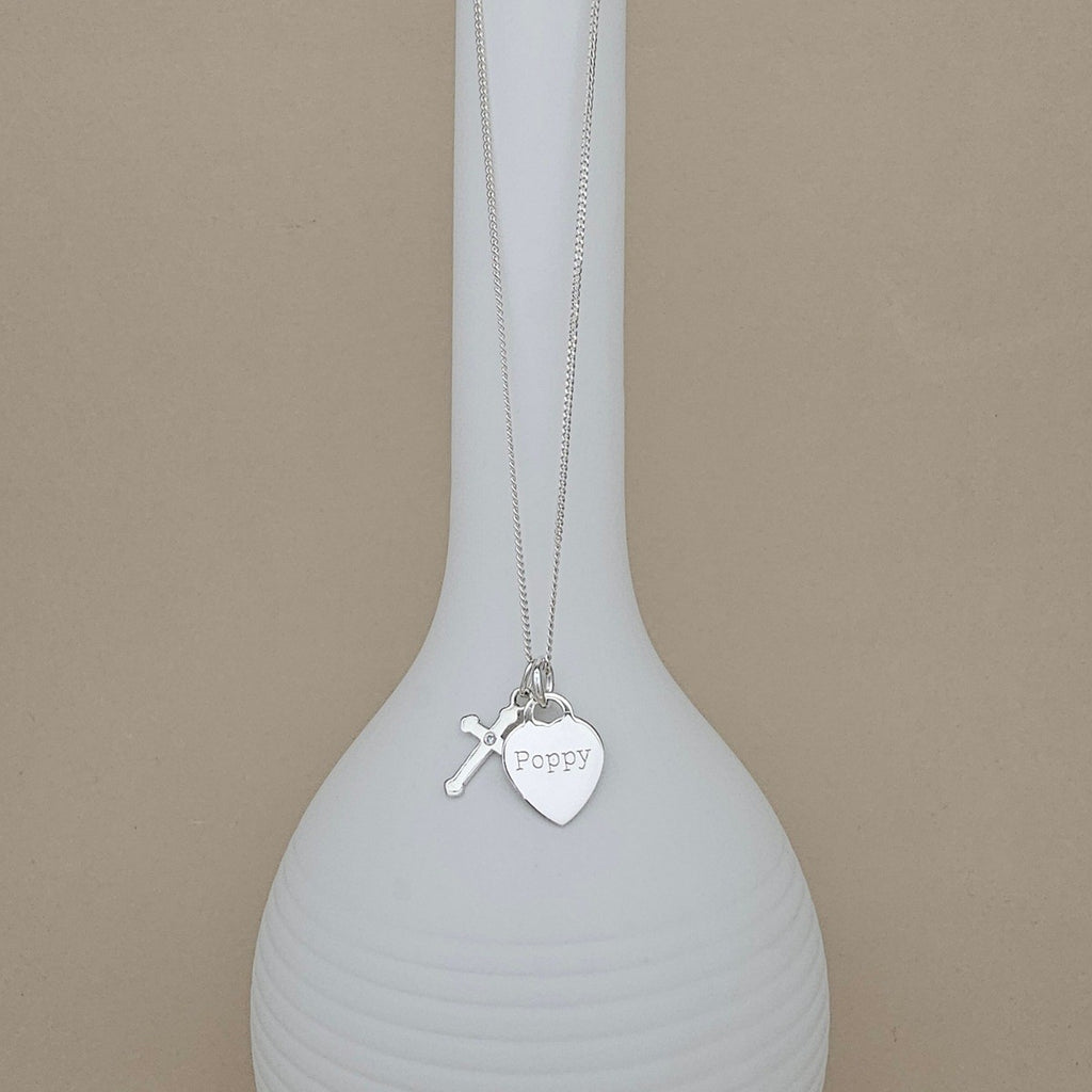 A silver necklace with a small cross charm and a heart-shaped pendant engraved with the name 'Poppy', displayed draped over a white ribbed vase against a beige background.
