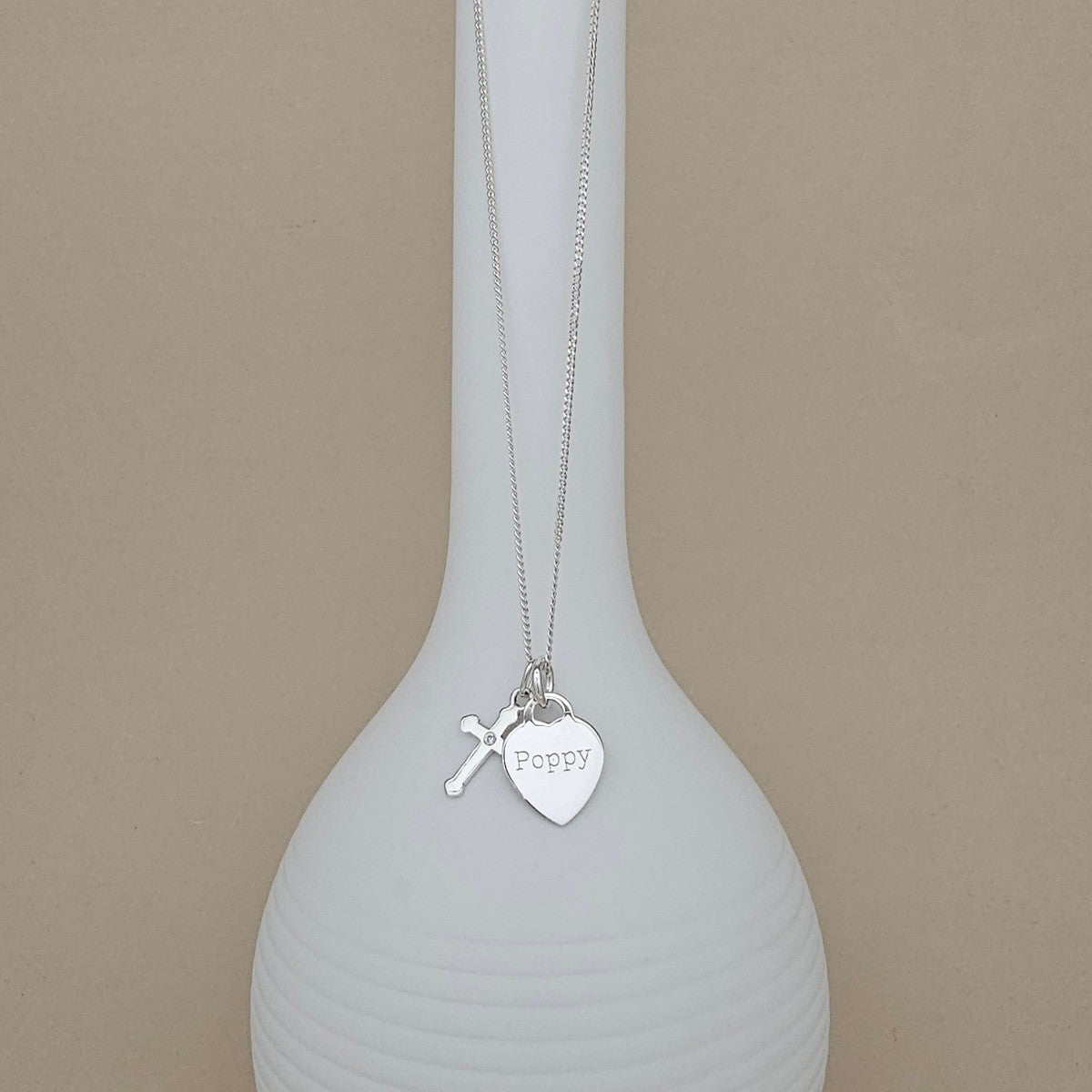A silver necklace with a small cross charm and a heart-shaped pendant engraved with the name 'Poppy', displayed draped over a white ribbed vase against a beige background.