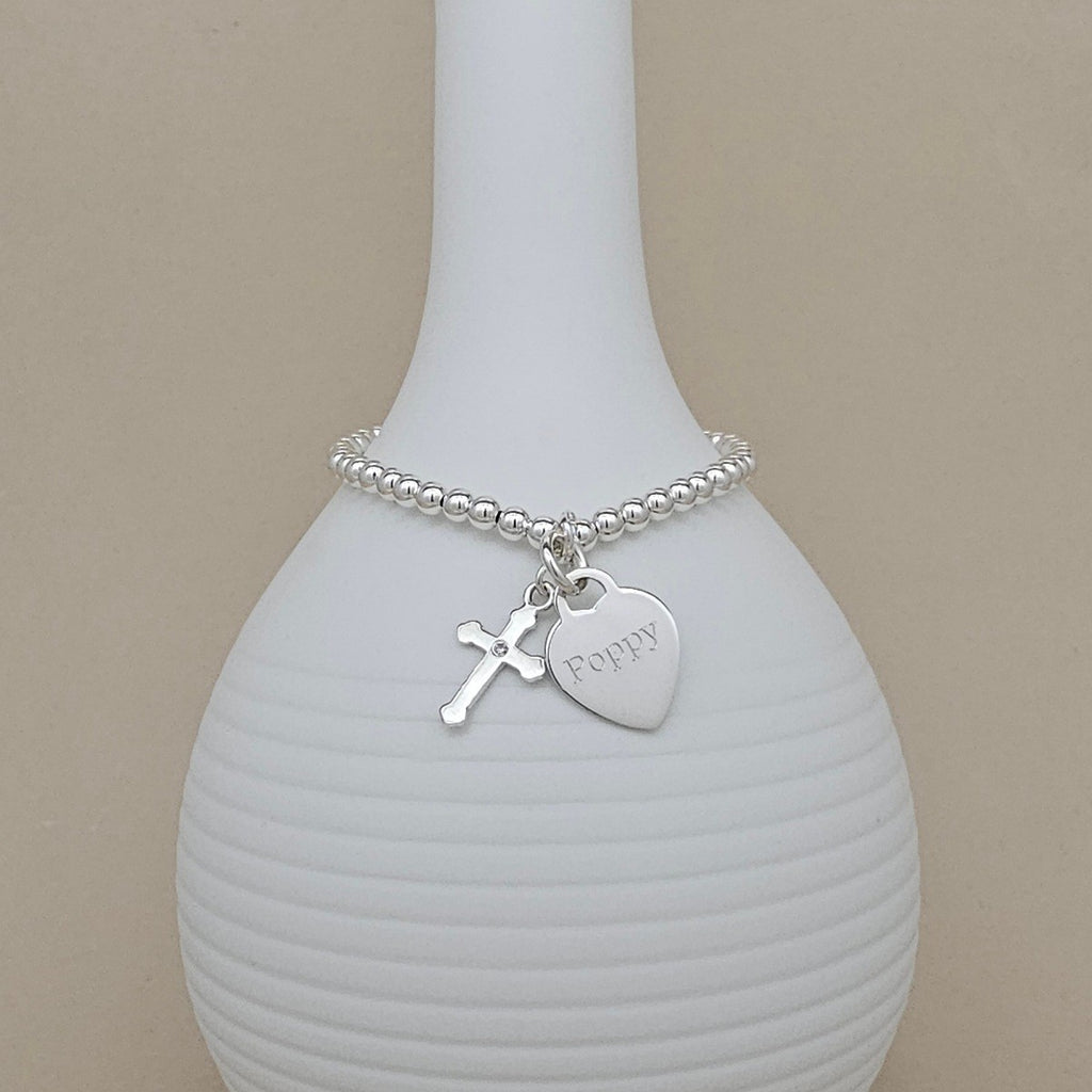 A silver beaded bracelet with a cross charm and a heart-shaped 'Poppy' engraving, displayed on a ribbed white ceramic vase against a beige background.