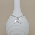 A silver beaded bracelet with a cross charm and a heart-shaped 'Poppy' engraving, displayed on a ribbed white ceramic vase against a beige background.