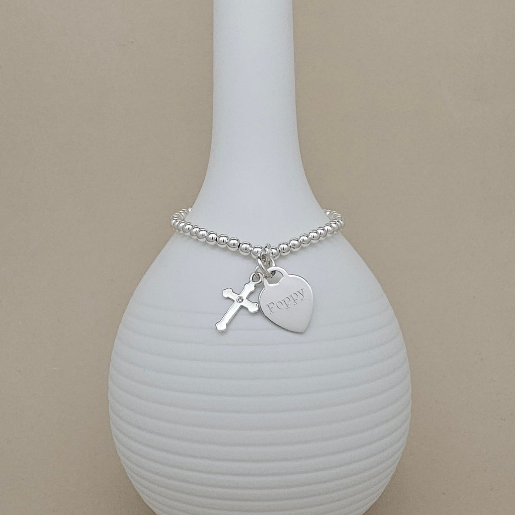 A silver beaded bracelet with a cross charm and a heart charm engraved with the name 'Poppy', displayed on a white ribbed vase against a beige background.