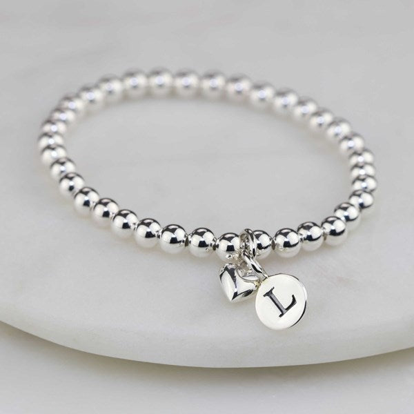 Silver beaded bracelet with a heart charm and round initial charm on a light gray background