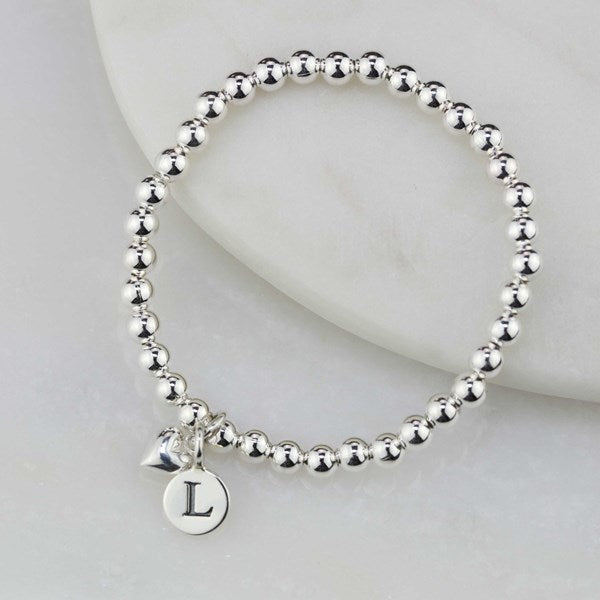 A silver beaded bracelet with a small initial 'L' charm and a tiny heart charm, displayed on a white marble prop.