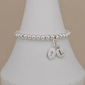 A silver beaded stretch bracelet with two oval charms, one with a cross and the other with the letter 'L', displayed on a white cone stand against a beige background.