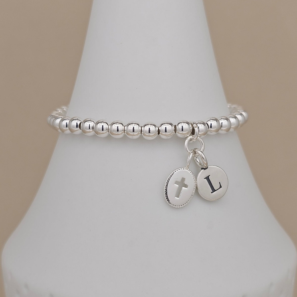 A silver beaded stretch bracelet with two oval charms, one with a cross and the other with the letter 'L', displayed on a white cone stand against a beige background.