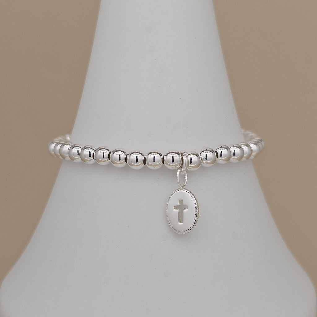 A silver beaded bracelet with a small oval charm featuring a cross, displayed on a white cone-shaped jewelry stand against a warm beige background.