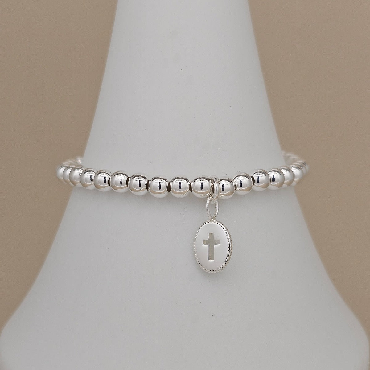 A silver beaded bracelet with a small oval charm featuring a cross, displayed on a white cone-shaped jewelry stand against a warm beige background.