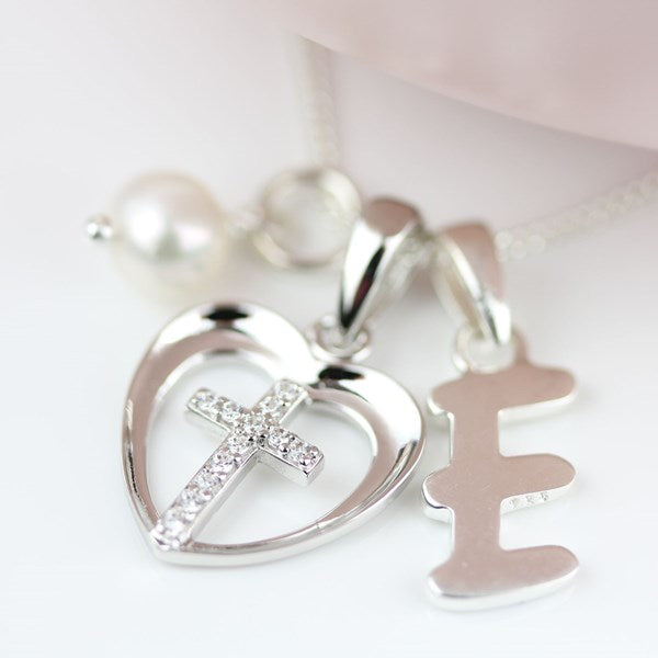 Silver heart-shaped pendant necklace with a cross studded with small clear crystals, alongside a silver letter 'E' pendant and a pearl bead on a white background.