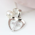 Silver heart-shaped pendant necklace with an open heart design and a small cross in the center, embellished with clear rhinestones. A white pearl bead is attached near the top, and a silver chain is visible against a soft white background.