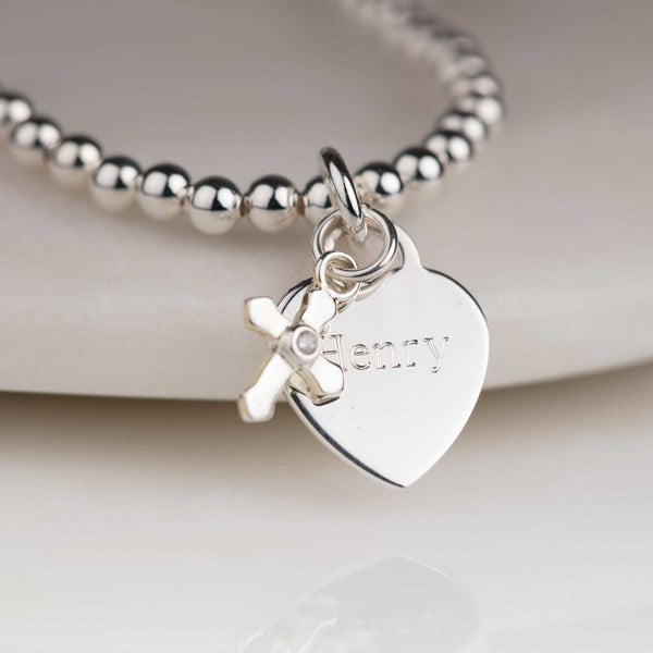 Silver bracelet with heart-shaped tag and cross charm on a light background