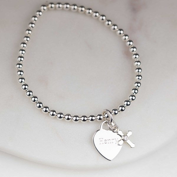 A silver beaded bracelet with a heart-shaped charm engraved with the name 'Henry' and a small cross charm, displayed on a light marble surface.