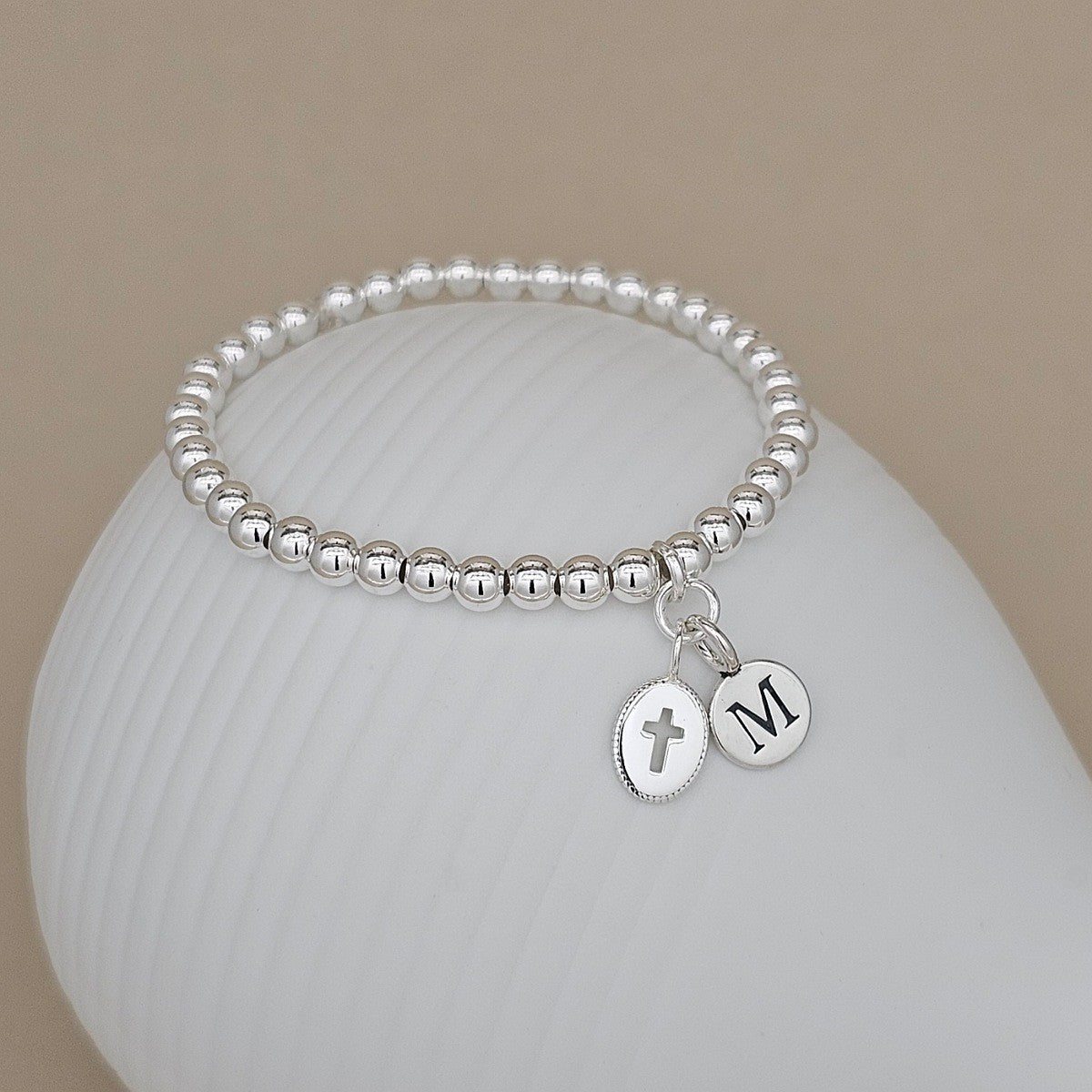 A silver beaded stretch bracelet with two oval charms, one with a cross and the other with the letter 'M', displayed on a white ribbed ceramic stand against a beige background.