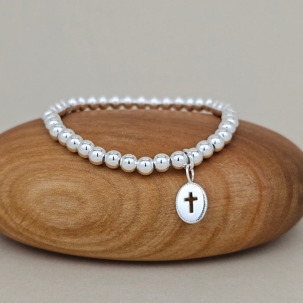A silver beaded stretch bracelet with a small oval silver pendant featuring a cross, displayed on a smooth wooden stone against a neutral beige background.