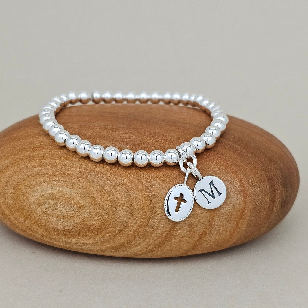 Silver beaded bracelet with a cross and letter 'M' charm on a wooden surface