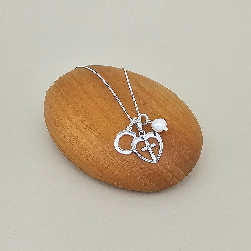 A silver necklace with two interlocking heart charms, one featuring a small cross, displayed on a smooth wooden block against a light beige background.