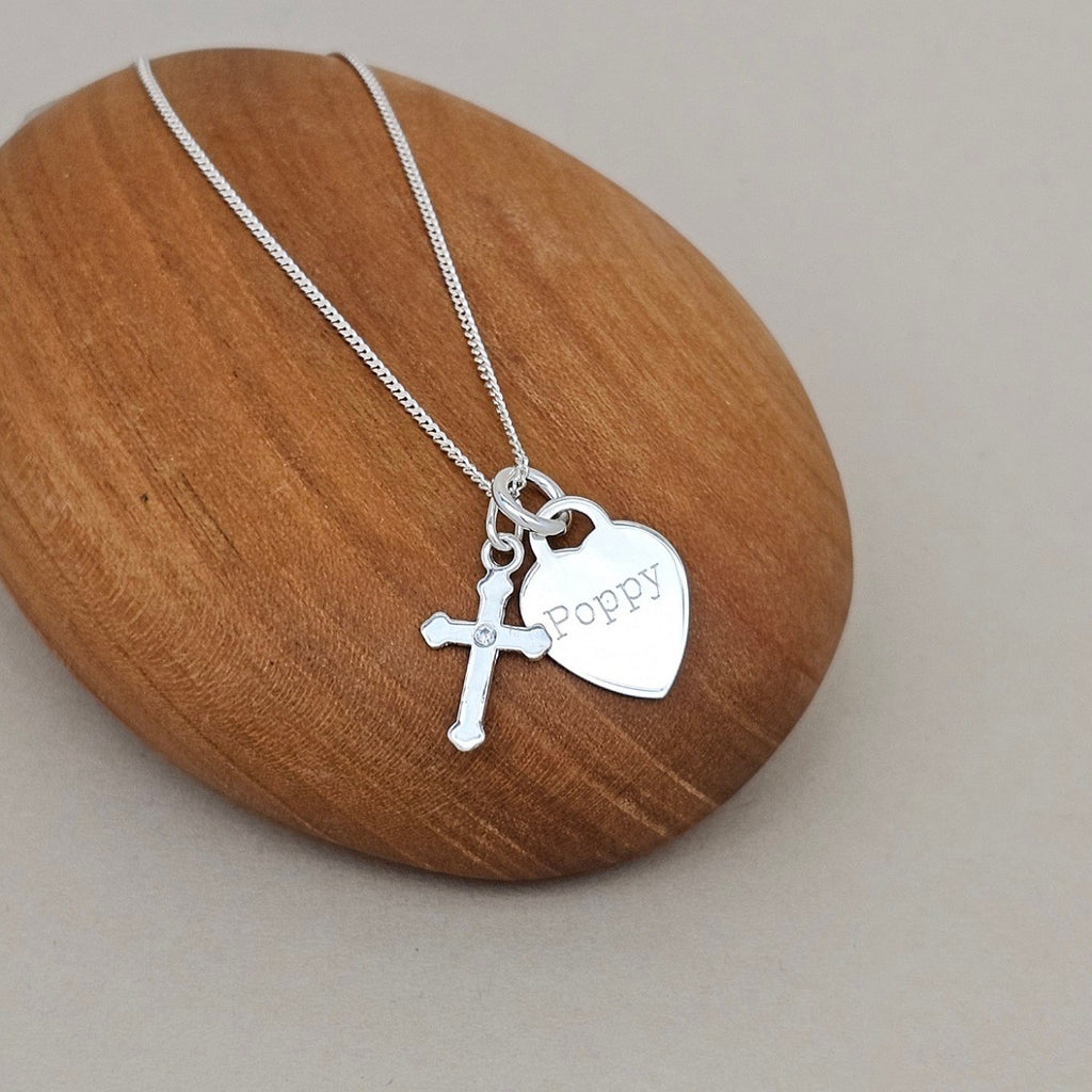 A silver cross and heart pendant necklace with the name 'Poppy' engraved on the heart, displayed on a smooth wooden block against a neutral gray background.