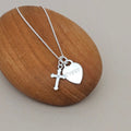 A silver cross and heart pendant necklace with the name 'Poppy' engraved on the heart, displayed on a smooth wooden block against a neutral gray background.
