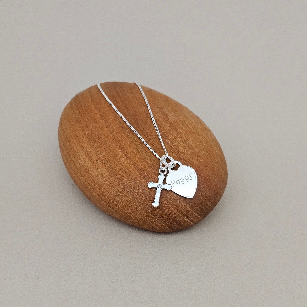 A silver necklace with a cross and a heart-shaped charm engraved with 'happy', displayed on a smooth, oval wooden stand against a neutral gray background.