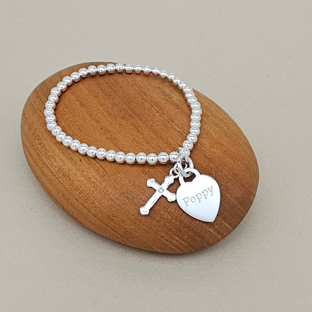 A silver beaded bracelet with a cross charm and a heart-shaped tag engraved with the name 'Poppy', displayed on a smooth wooden stone against a neutral beige background.