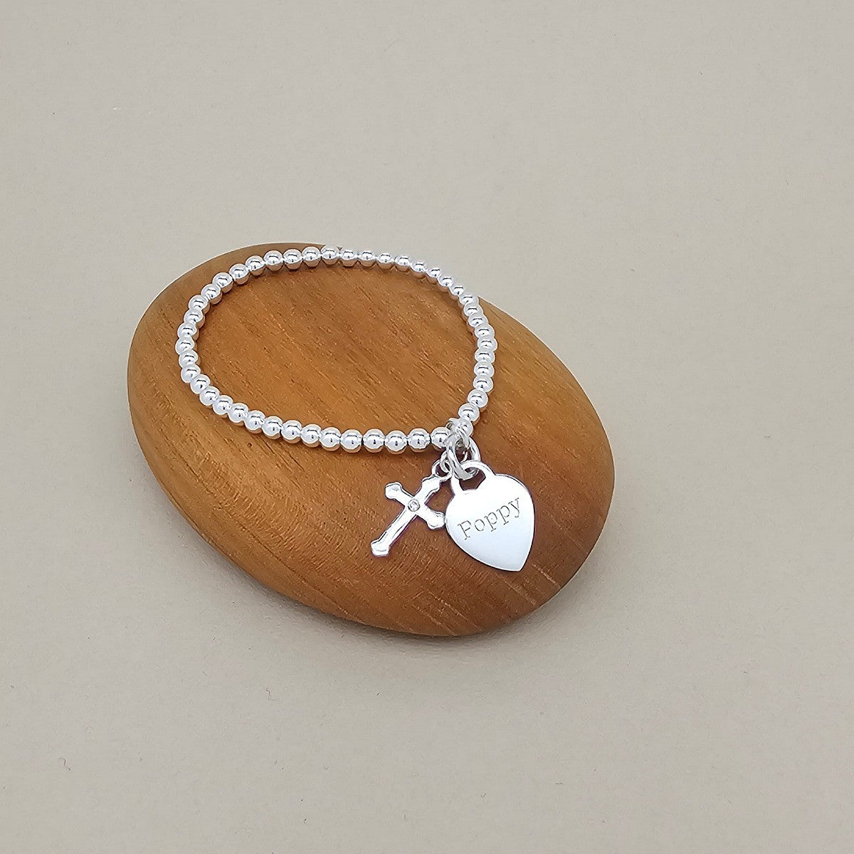 A silver beaded bracelet with a cross charm and a heart-shaped 'Poppy' pendant, displayed on a smooth wooden stone against a neutral beige background.