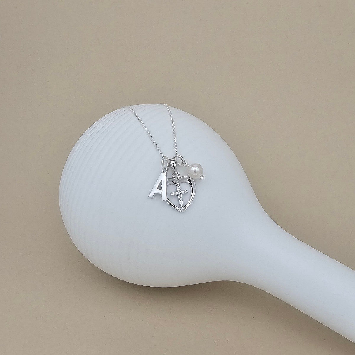 A silver necklace with a heart-shaped pendant, a small pearl bead, and a letter 'A' initial charm, displayed on a white ribbed spoon against a beige background.