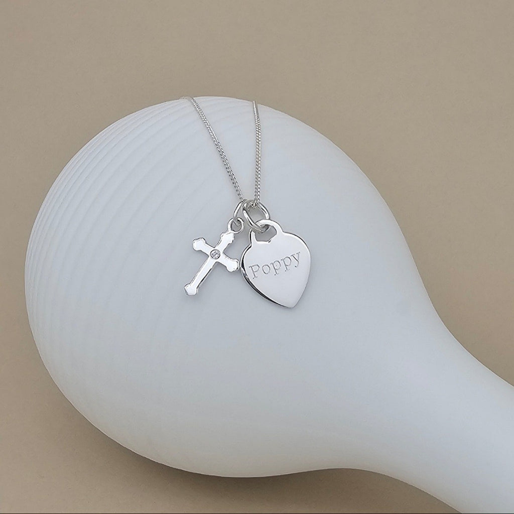 A silver necklace with a heart-shaped charm engraved with the name 'Poppy' and a small cross charm, displayed on a white ceramic surface against a warm beige background.