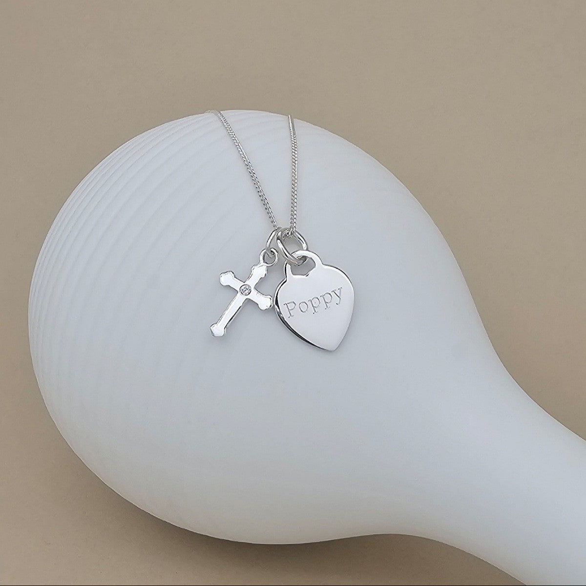 A silver necklace with a heart-shaped charm engraved with the name 'Poppy' and a small cross charm, displayed on a white ceramic surface against a warm beige background.