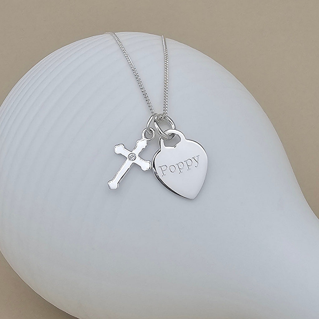A silver necklace with a heart-shaped charm engraved with the name 'Poppy' and a small cross charm, displayed on a white ceramic surface against a warm beige background.