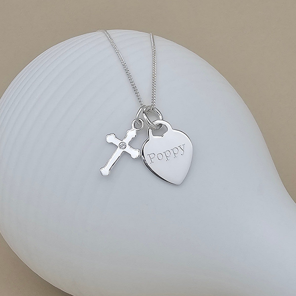A silver necklace with a heart-shaped charm engraved with the name 'Poppy' and a small cross charm, displayed on a white ceramic surface against a warm beige background.