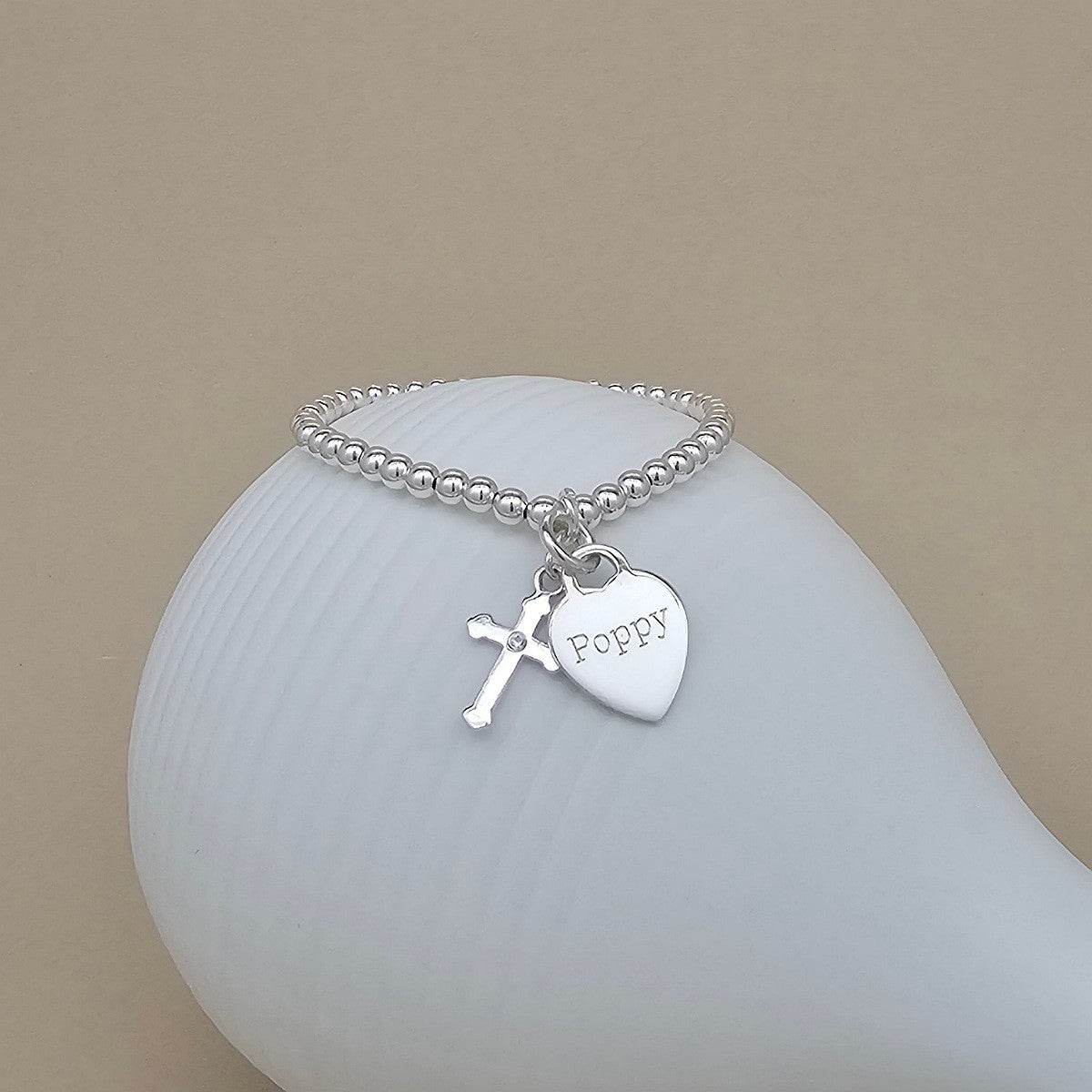 A silver beaded bracelet with a small cross charm and a heart-shaped charm engraved with the name 'Poppy', displayed on a white ceramic stand against a beige background.