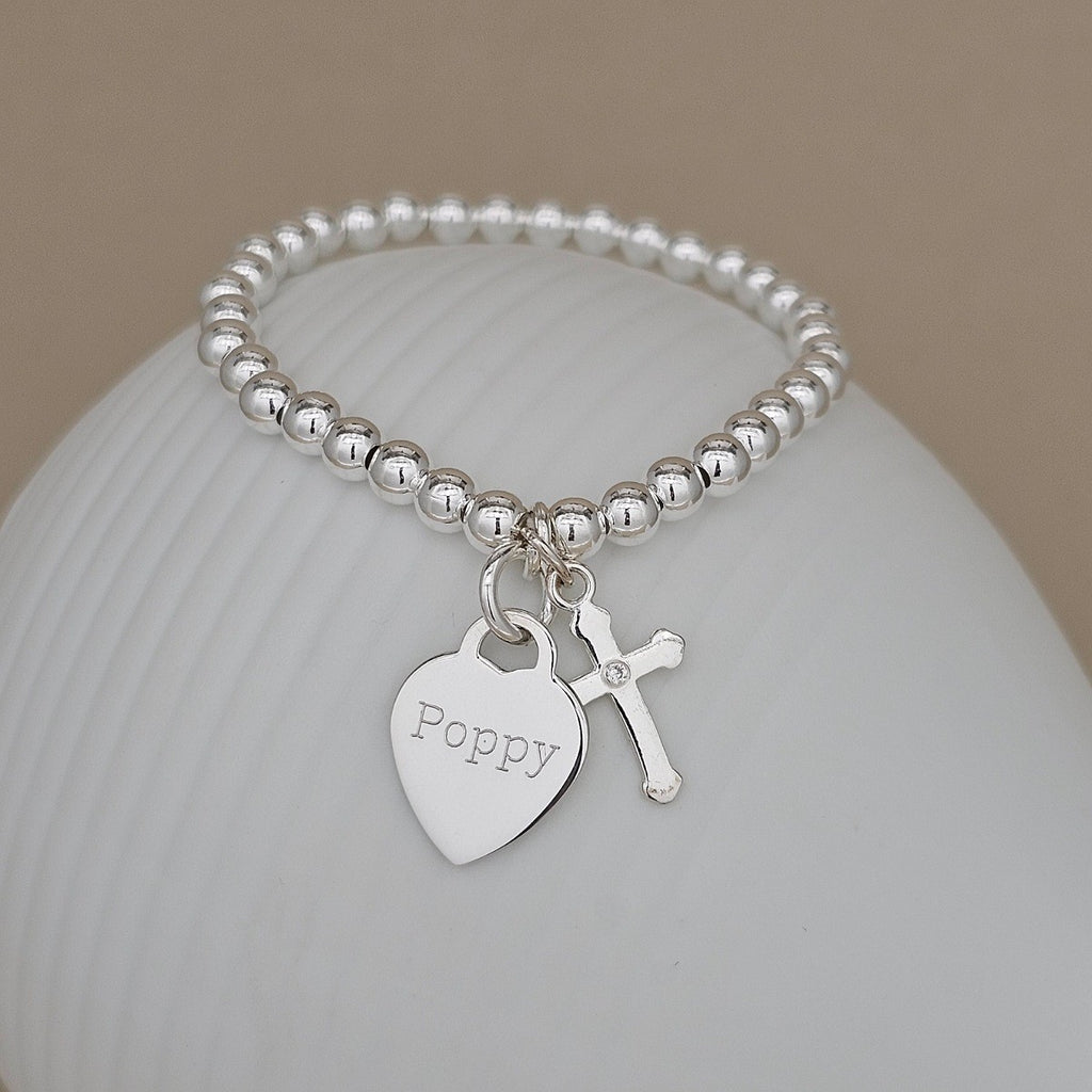 A silver beaded bracelet with a heart-shaped charm engraved with the name 'Poppy' and a small cross charm, displayed on a white ceramic surface against a warm beige background.