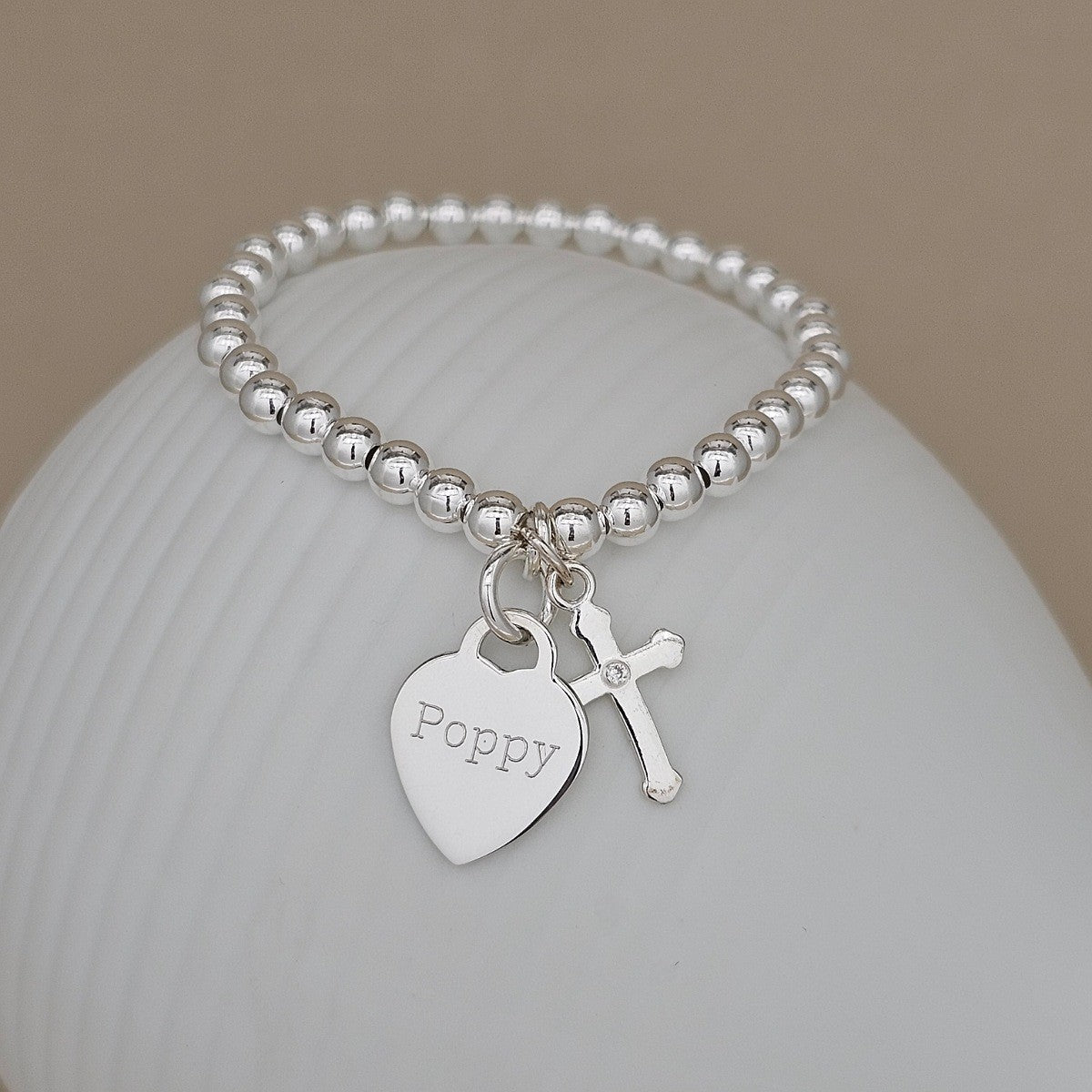 A silver beaded bracelet with a heart-shaped charm engraved with the name 'Poppy' and a small cross charm, displayed on a white ceramic surface against a warm beige background.