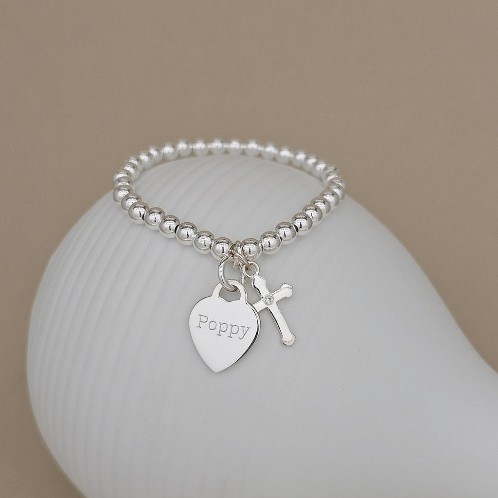 A silver beaded bracelet with a heart-shaped charm engraved with the name 'Poppy' and a small cross charm, displayed on a white ceramic surface against a warm beige background.