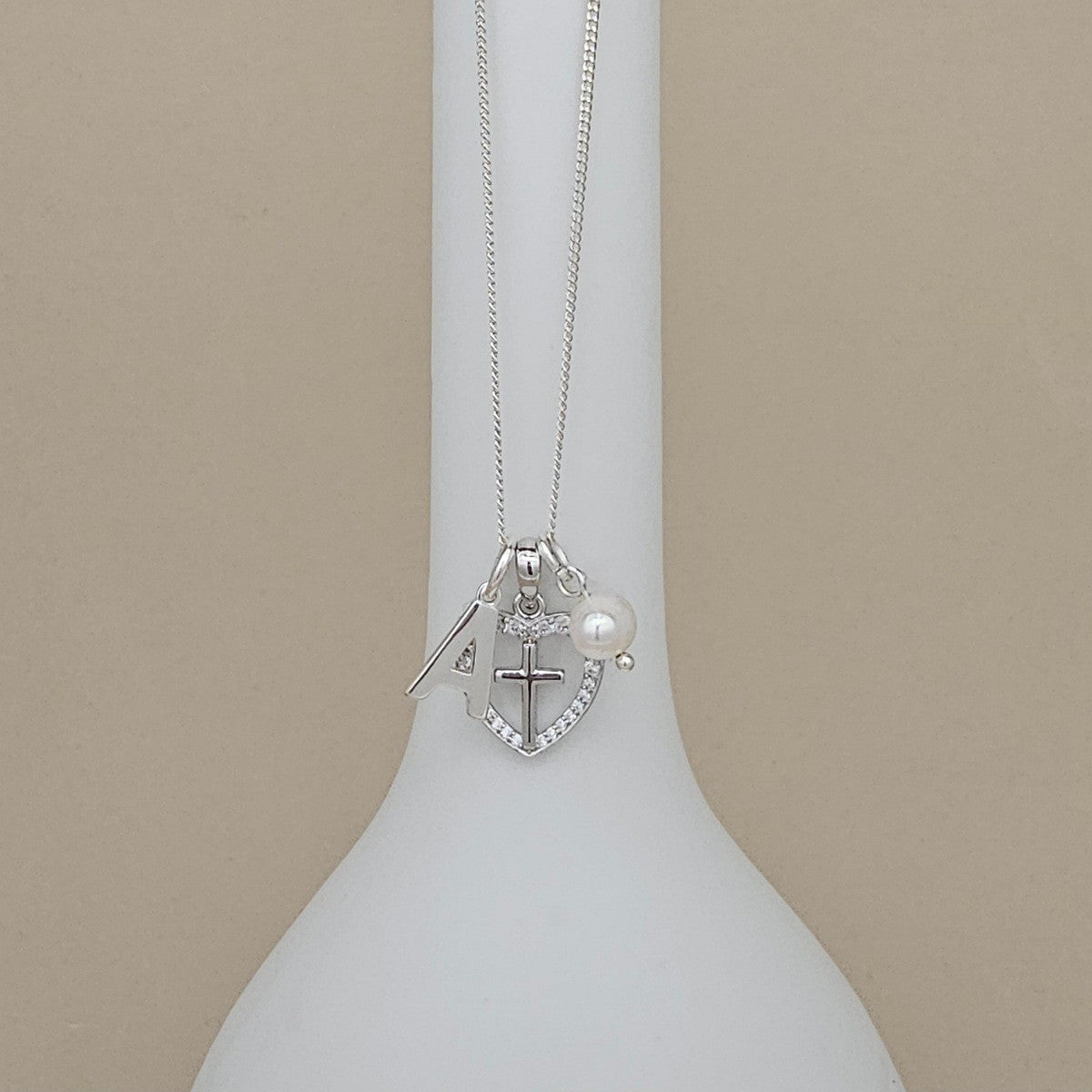 Silver necklace with a cross and heart pendant on a white stand against a beige background