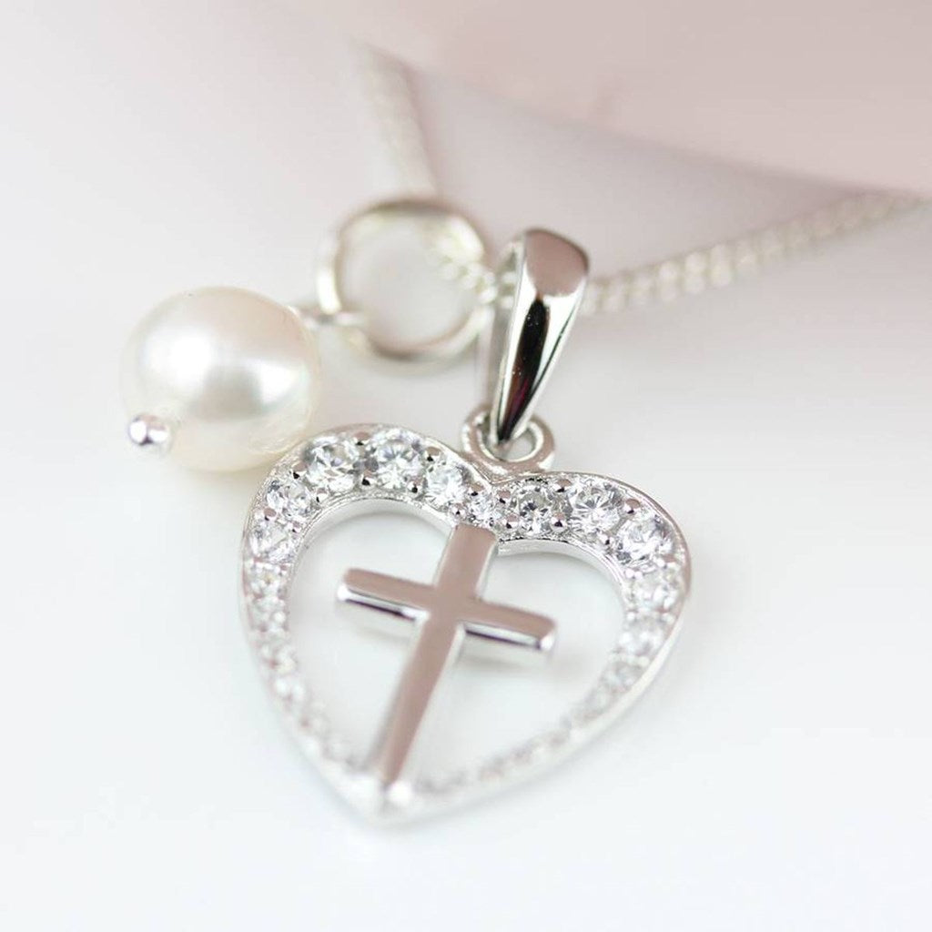 Silver heart-shaped pendant necklace with a cross in the center, surrounded by clear rhinestones, with a dangling white pearl charm on a silver chain.