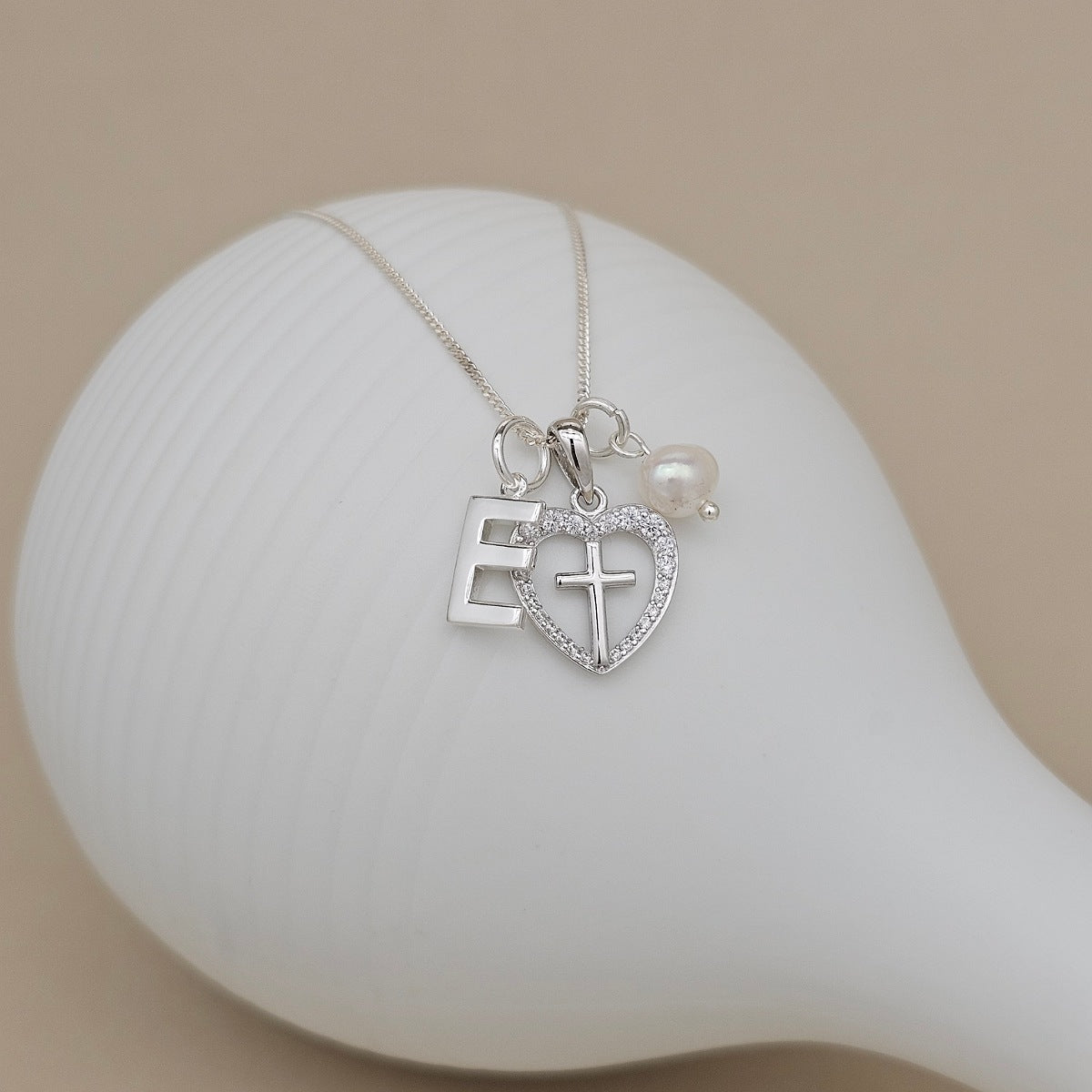 A silver initial necklace with a letter 'E' pendant, a heart-shaped crystal border with a cross in the center, and a small pearl charm, displayed on a white ceramic stand against a beige background.