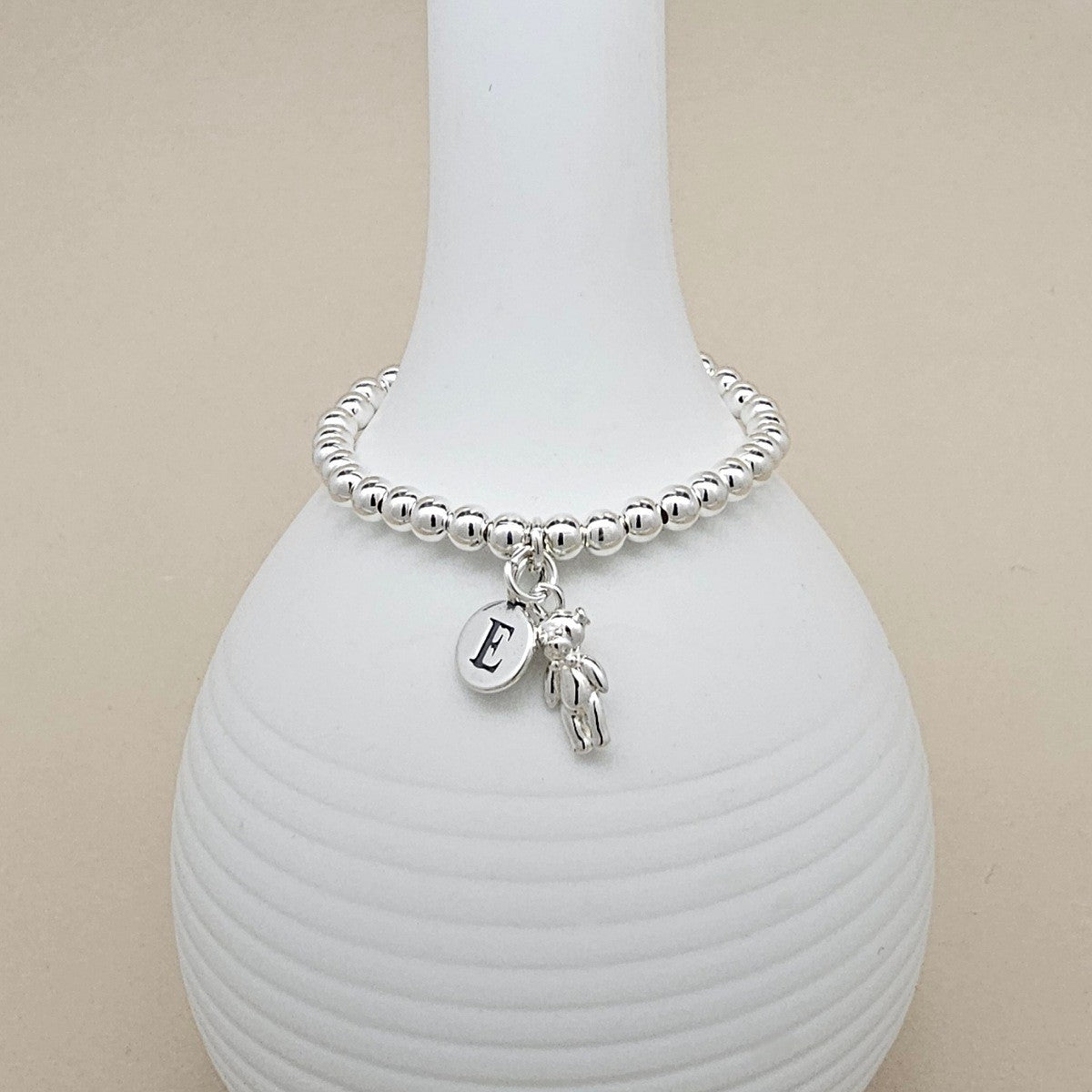 A silver beaded bracelet with a dangling initial 'E' charm and a small teddy bear charm, displayed on a white ribbed ceramic vase against a beige background.