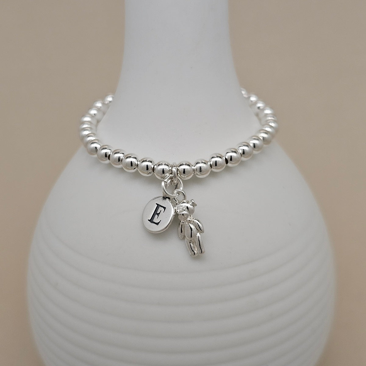 A silver beaded bracelet with a round initial 'E' charm and a small teddy bear charm, displayed on a white ceramic vase against a beige background.
