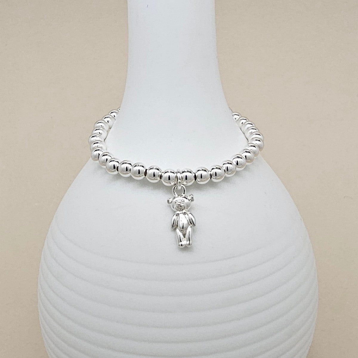 A silver beaded bracelet with a small teddy bear charm, displayed on a white ribbed ceramic vase against a light beige background.