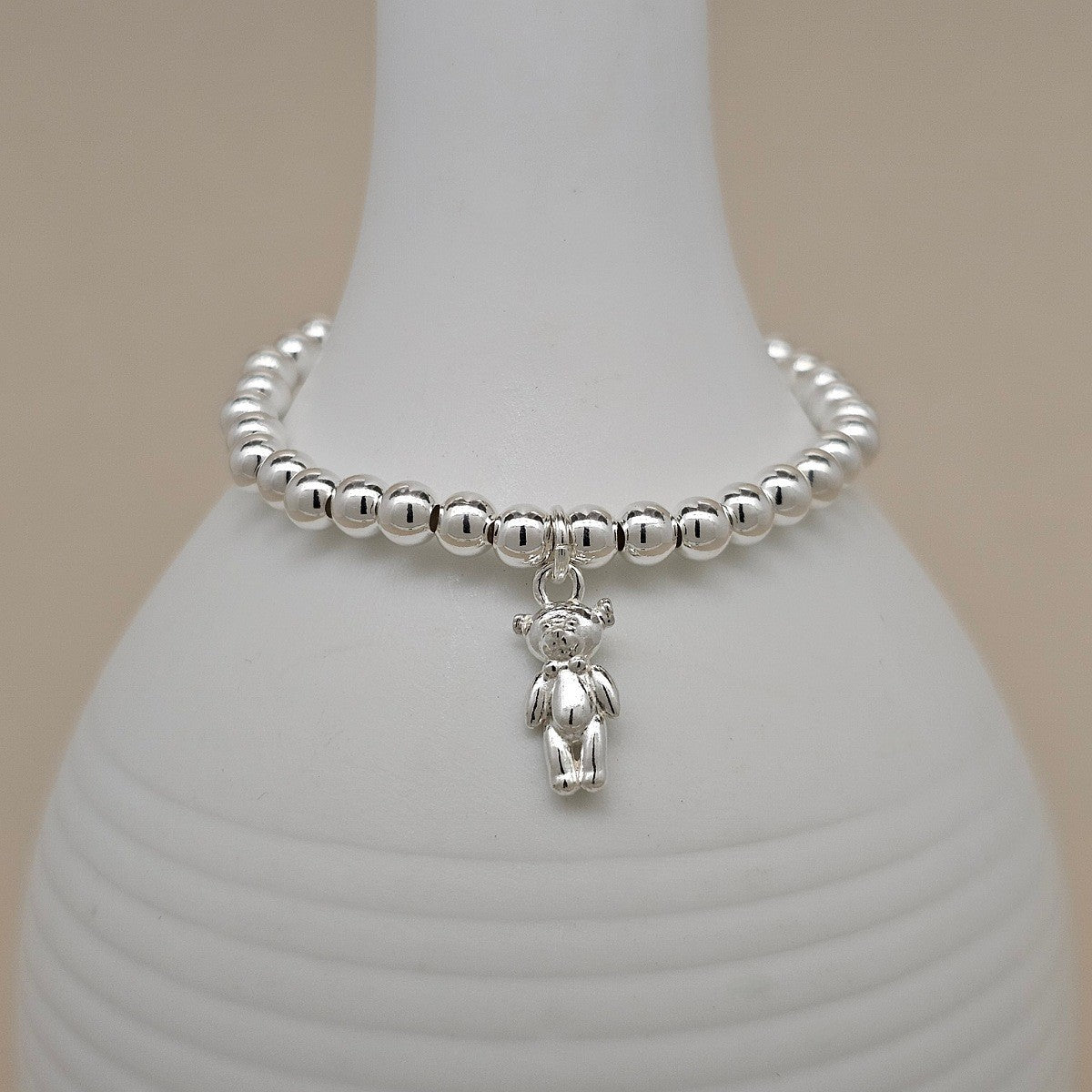 A silver beaded bracelet with a small teddy bear charm, displayed on a white ceramic vase against a warm beige background.