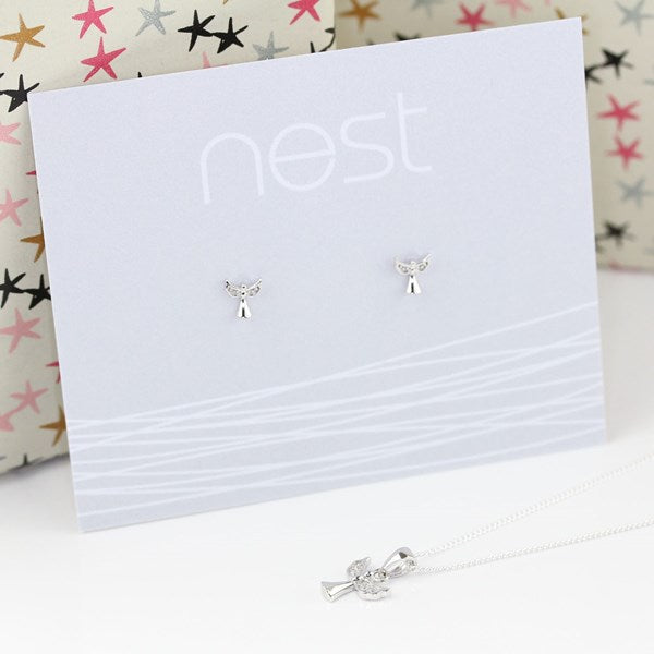 Sterling silver angel stud earrings on a white display card with the brand name 'nest', with a matching silver angel necklace in the foreground and a patterned gift box in the background.