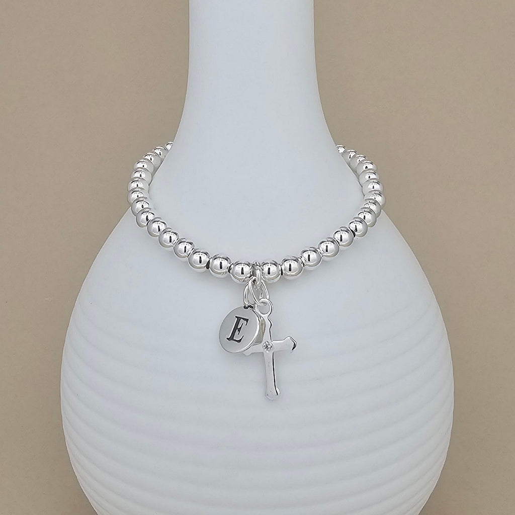 Silver beaded bracelet with a letter 'E' charm and cross pendant on a white vase.