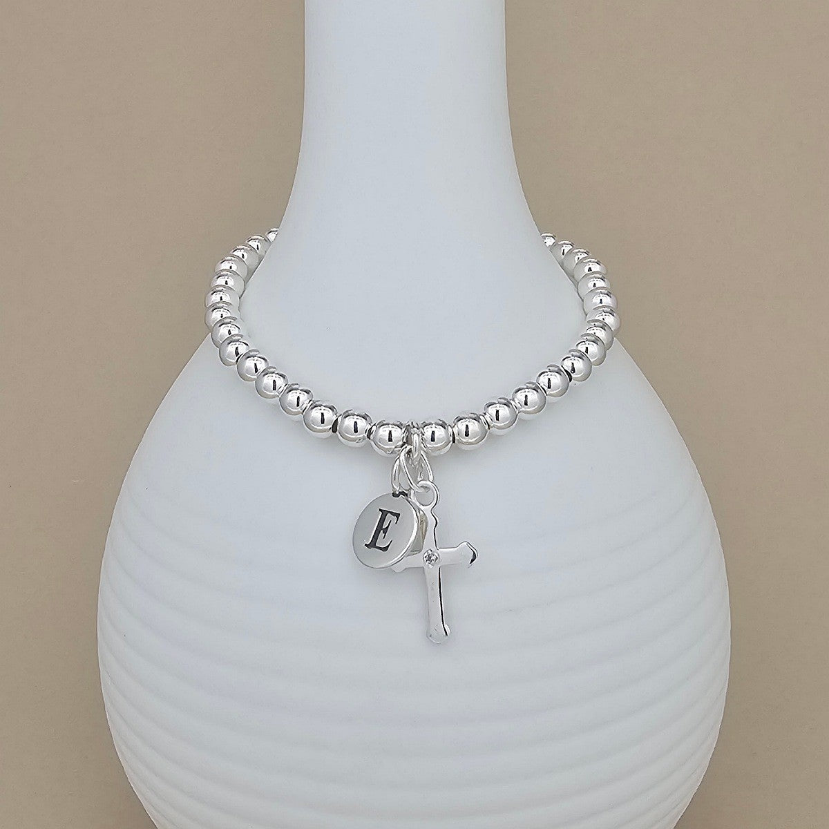 Silver beaded bracelet with a letter 'E' charm and cross pendant on a white vase.