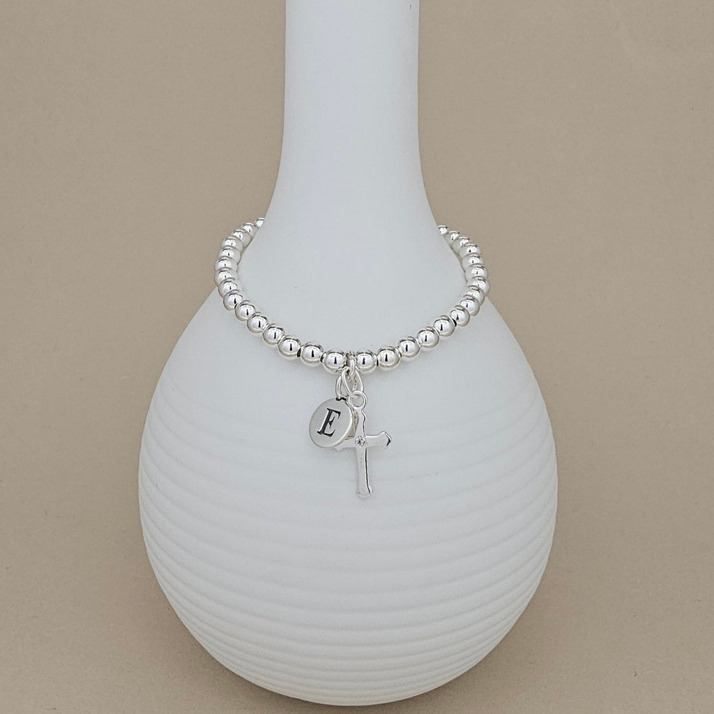 A silver beaded bracelet with a small initial 'E' charm and a cross pendant, displayed on a ribbed white ceramic vase against a solid beige background.