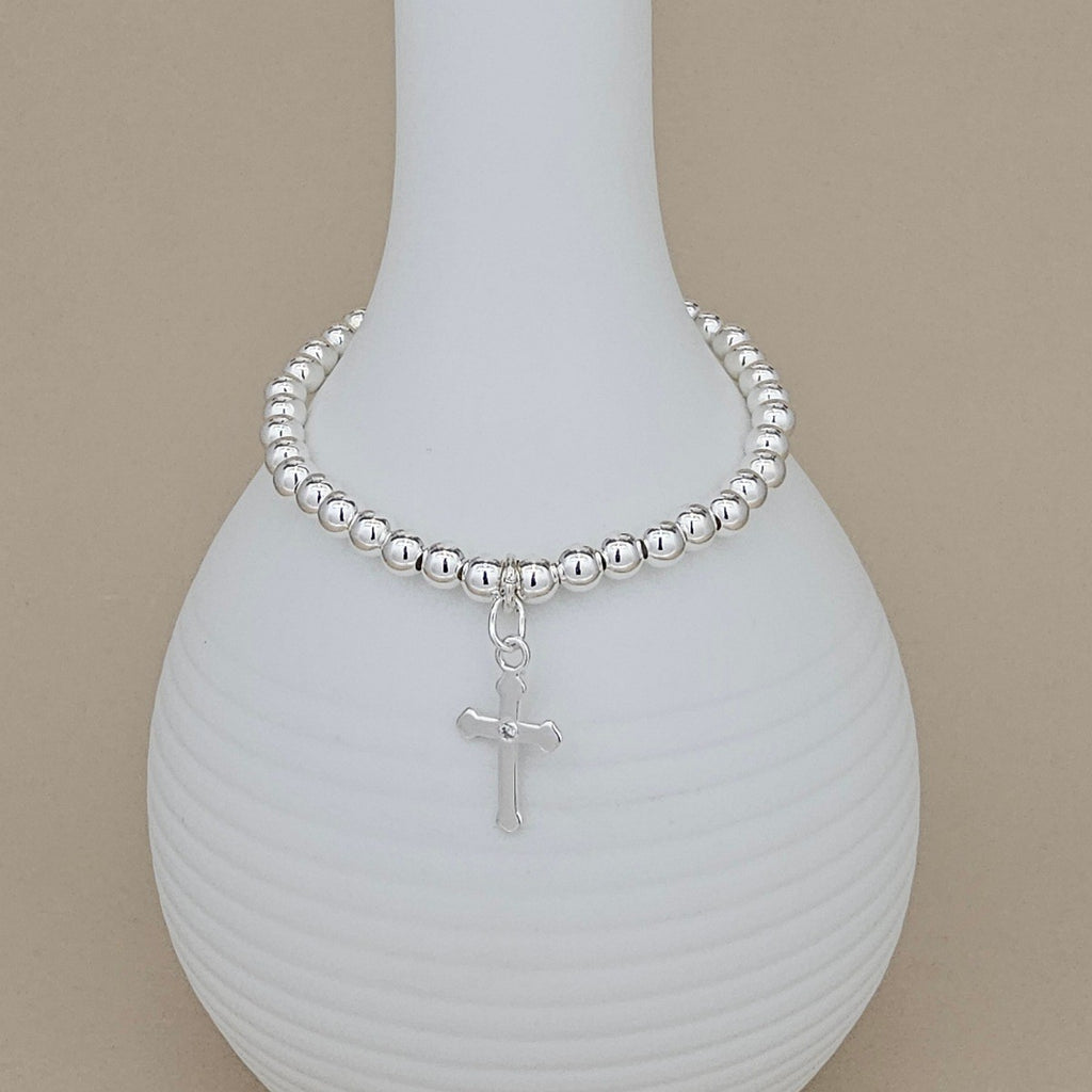 A silver beaded bracelet with a dangling cross charm, displayed on a white ribbed ceramic vase against a warm beige background.