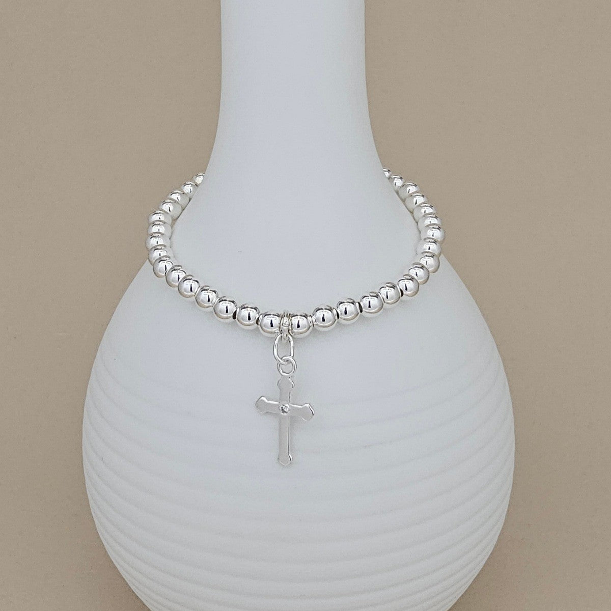 A silver beaded bracelet with a dangling cross charm, displayed on a white ribbed ceramic vase against a warm beige background.